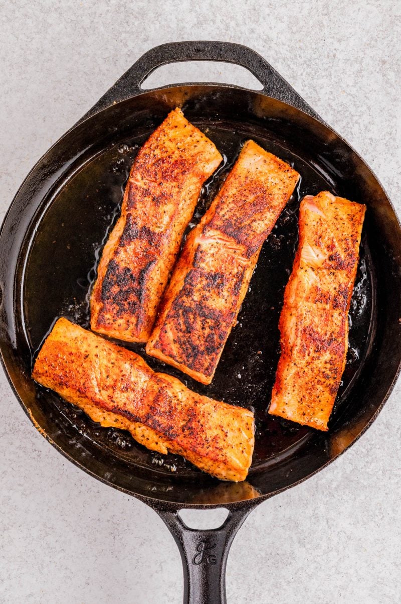 Four seasoned salmon fillets are searing in a black cast iron skillet on a light-colored surface.