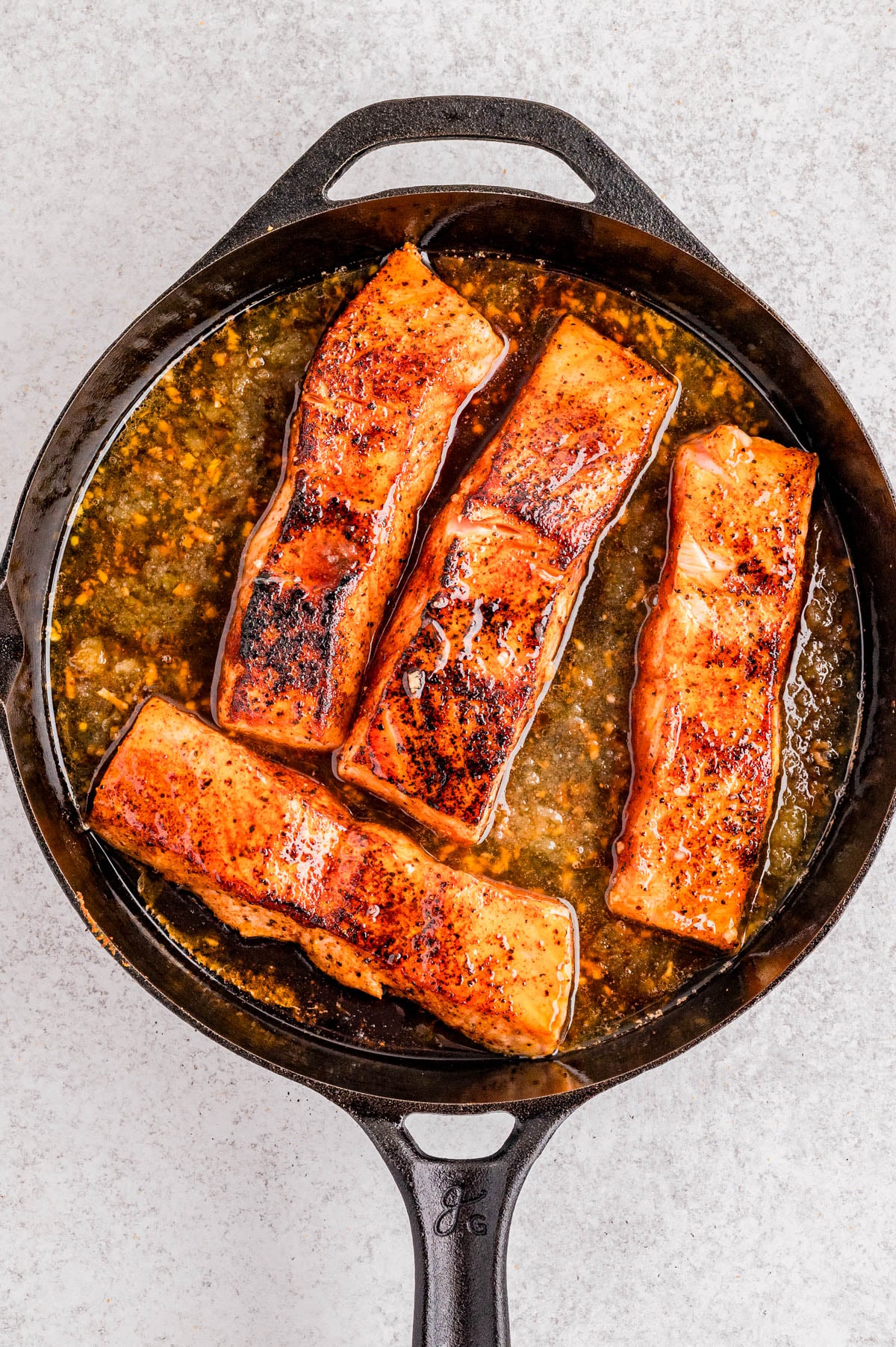 Four seared salmon fillets cooking in a cast iron skillet with a brown sauce on a light-colored surface.