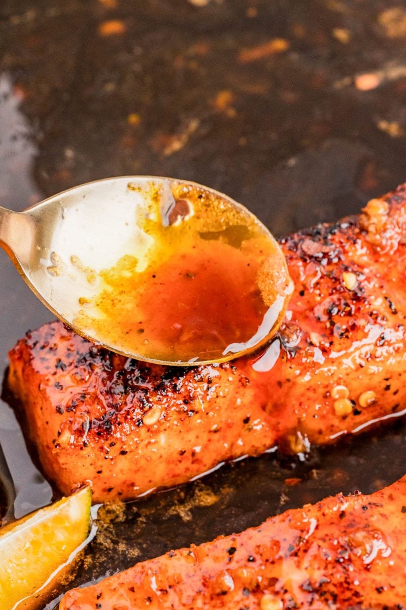 A spoon drizzles a brown sauce over a seasoned, cooked piece of salmon with visible spices, next to a lemon wedge.