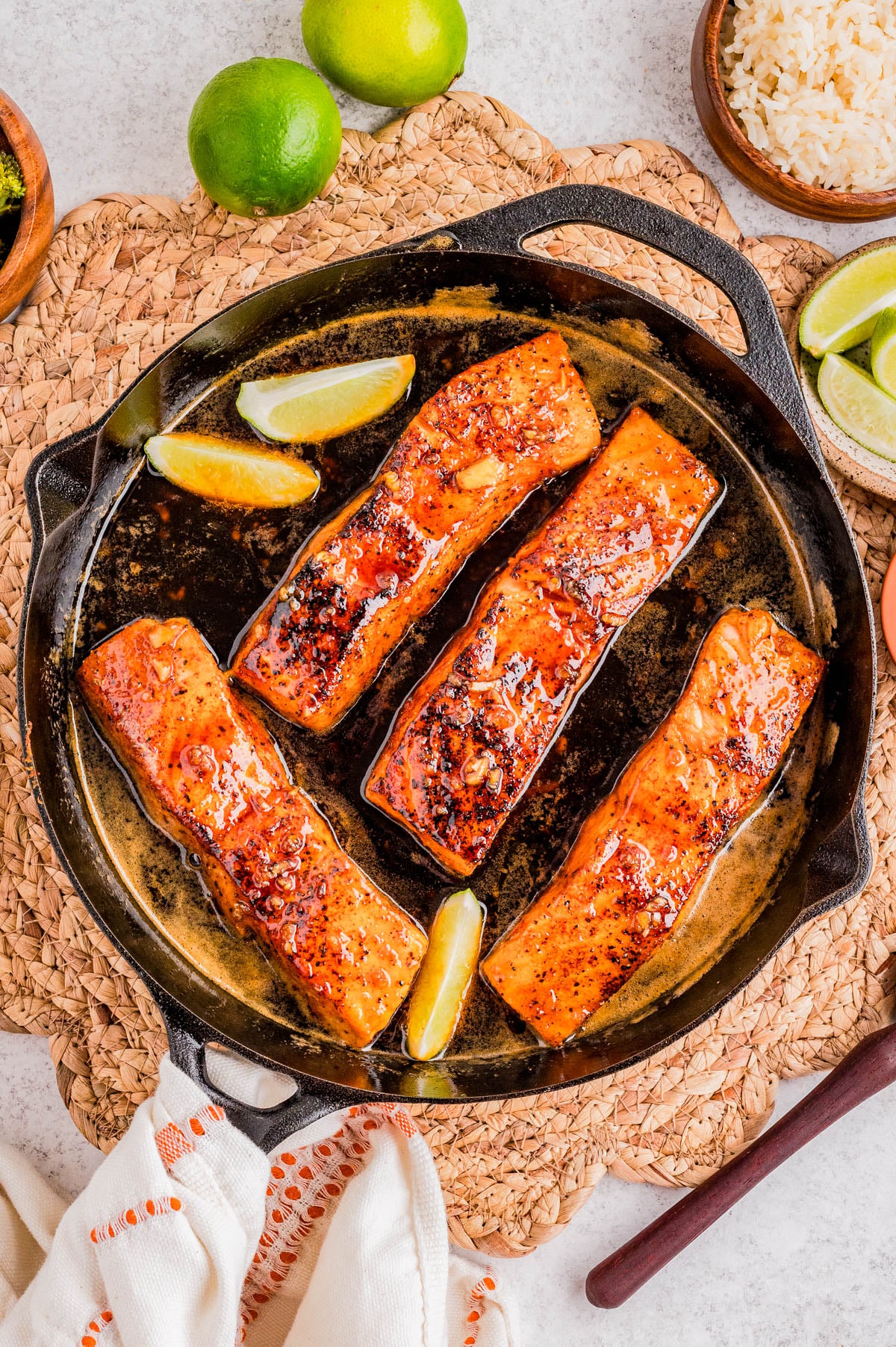 Four glazed salmon fillets with three lemon wedges in a cast iron skillet, surrounded by limes, rice, and a woven placemat.