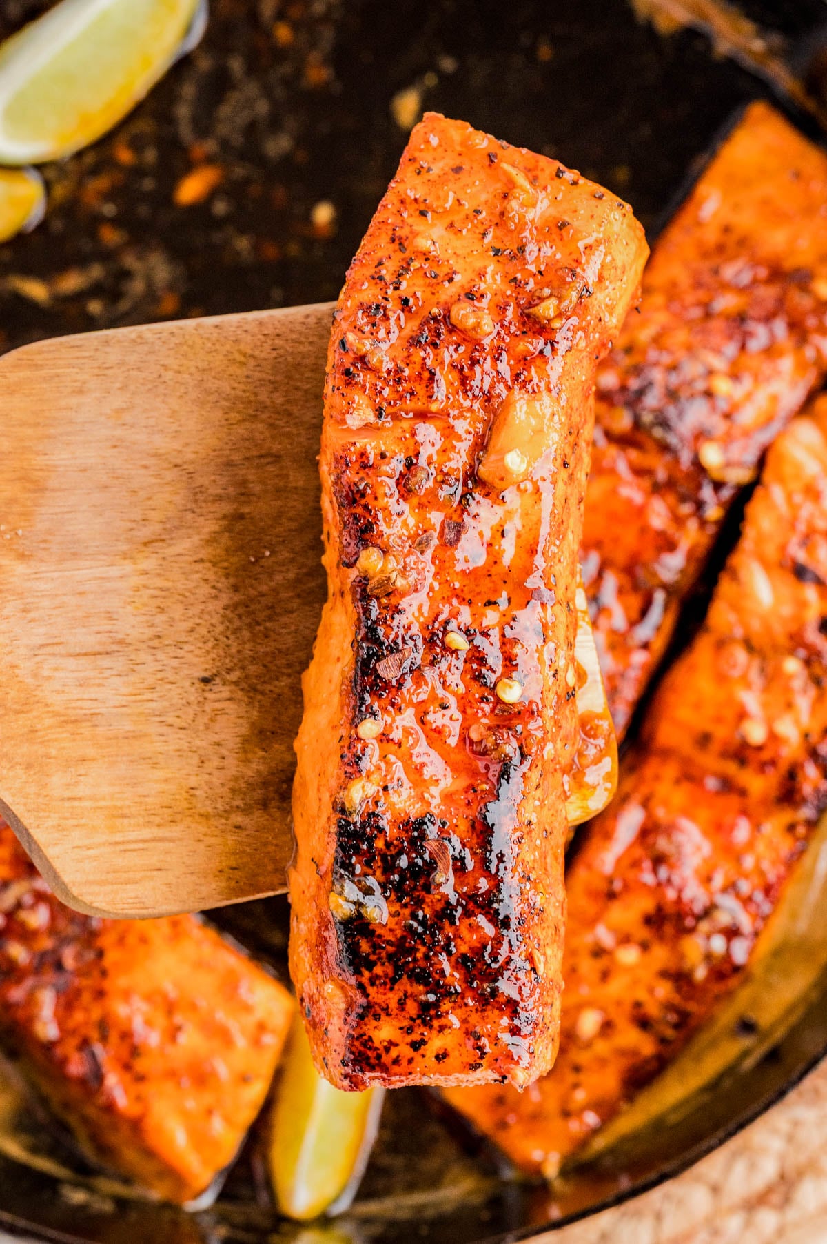 A cooked, seasoned salmon fillet is lifted from a pan with a wooden spatula, with other salmon pieces and lemon wedges visible in the background.