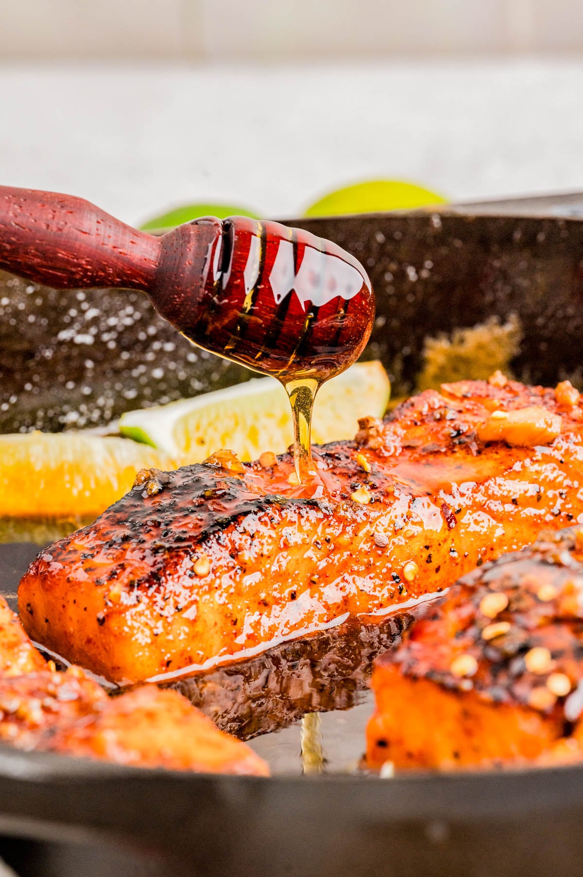 A hand drizzles honey over a seared, seasoned salmon fillet in a skillet, with lime wedges visible in the background.