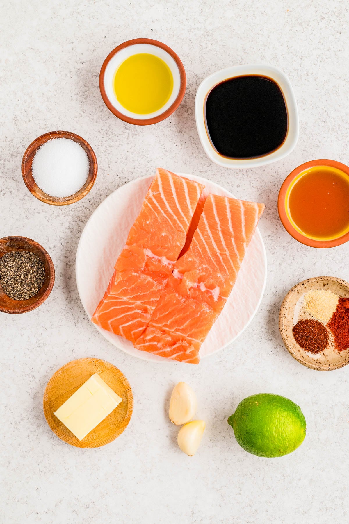 Two raw salmon fillets on a plate, surrounded by small bowls of olive oil, soy sauce, honey, salt, pepper, spices, butter, garlic cloves, and a whole lime on a white surface.