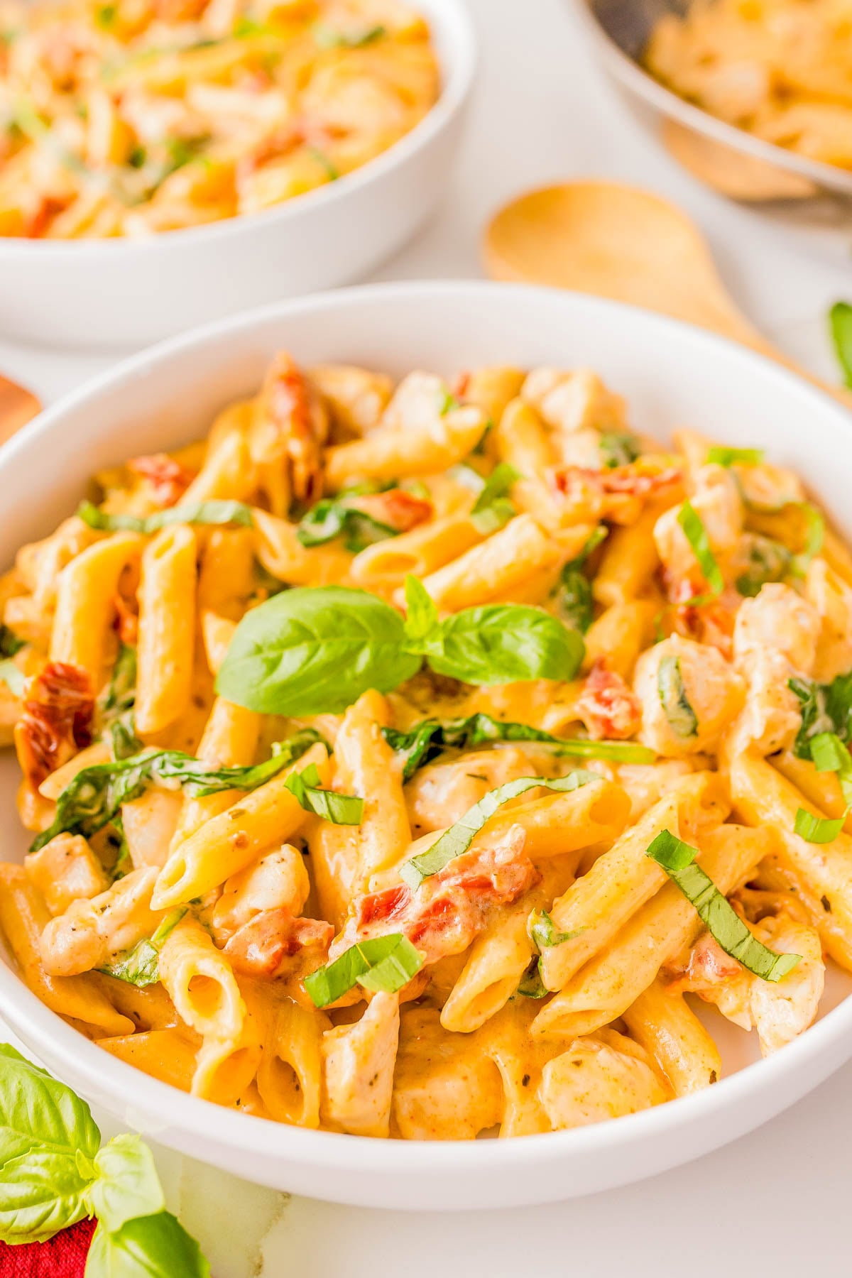 A white bowl filled with creamy penne pasta, pieces of chicken, sun-dried tomatoes, and fresh basil, with another pasta dish and wooden utensils in the background.