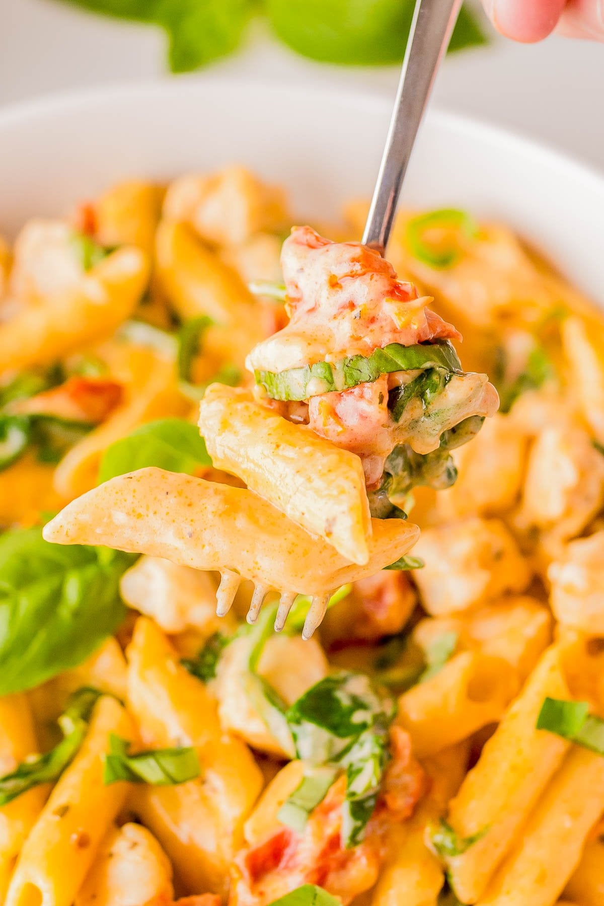 A close-up of a fork holding creamy penne pasta with pieces of chicken, sun-dried tomatoes, and fresh basil. A bowl of the same pasta is visible in the background.