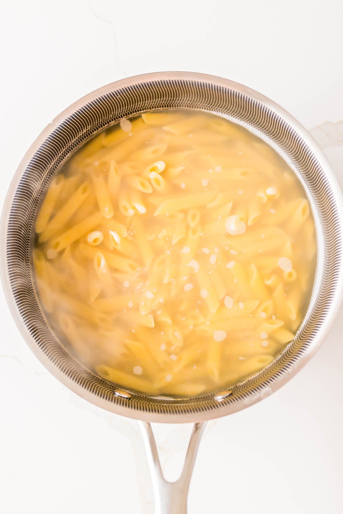 A pot of penne pasta boiling in water on a stovetop, viewed from above.