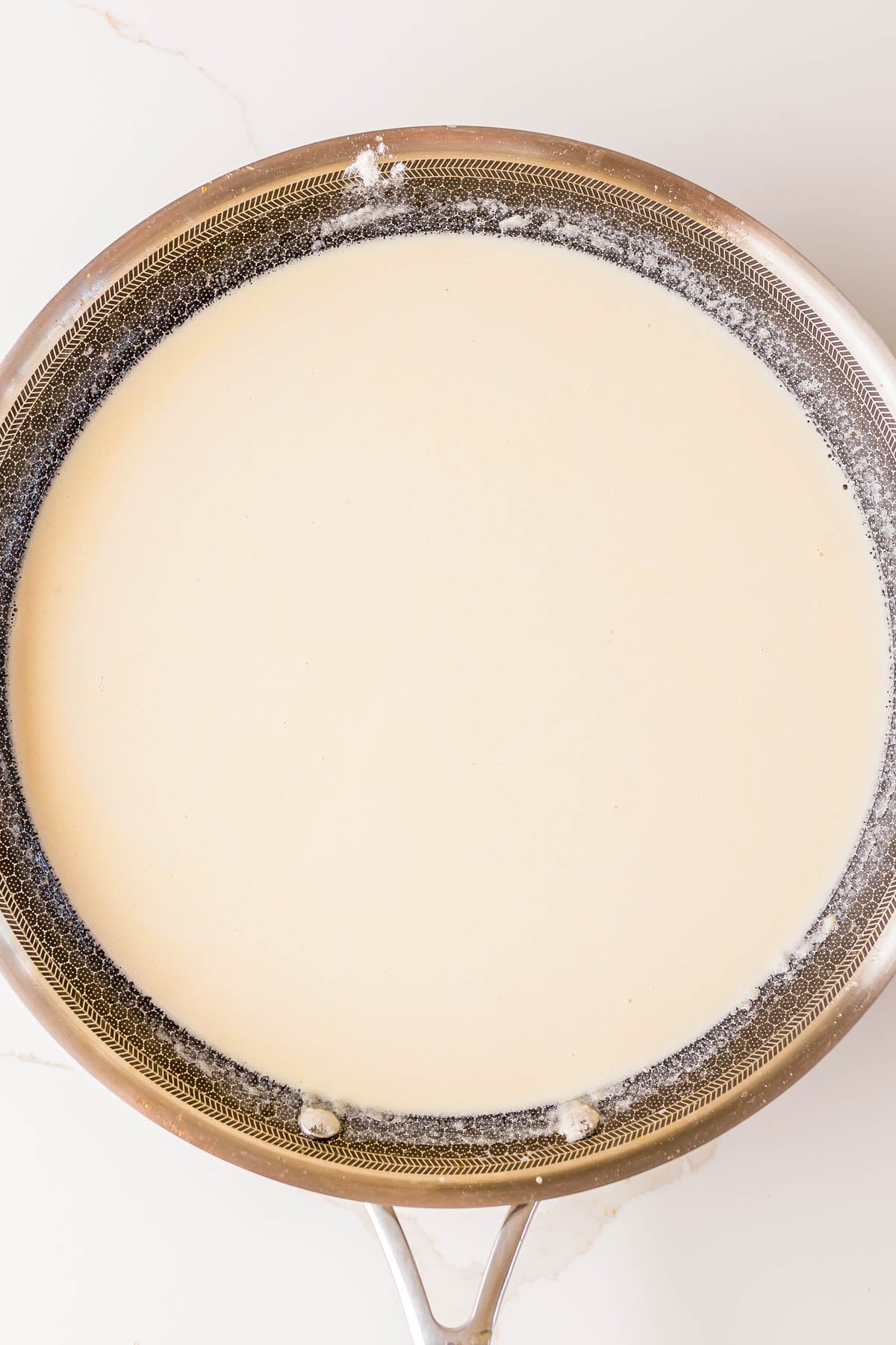 A stainless steel pan filled with a light-colored cream sauce, viewed from above on a white surface.