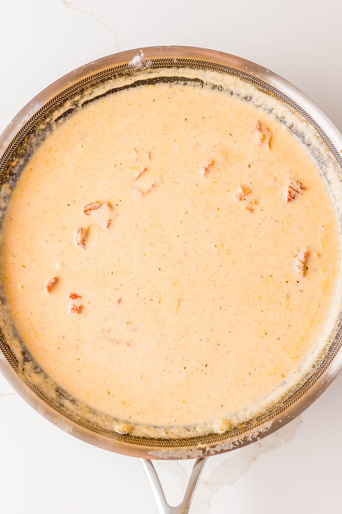 A stainless steel skillet filled with a creamy orange sauce with small pieces of tomato visible, sitting on a white countertop.