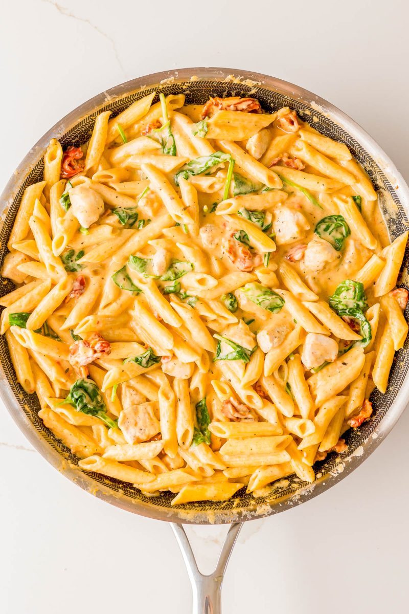 A skillet filled with creamy penne pasta, pieces of chicken, spinach, and sun-dried tomatoes.