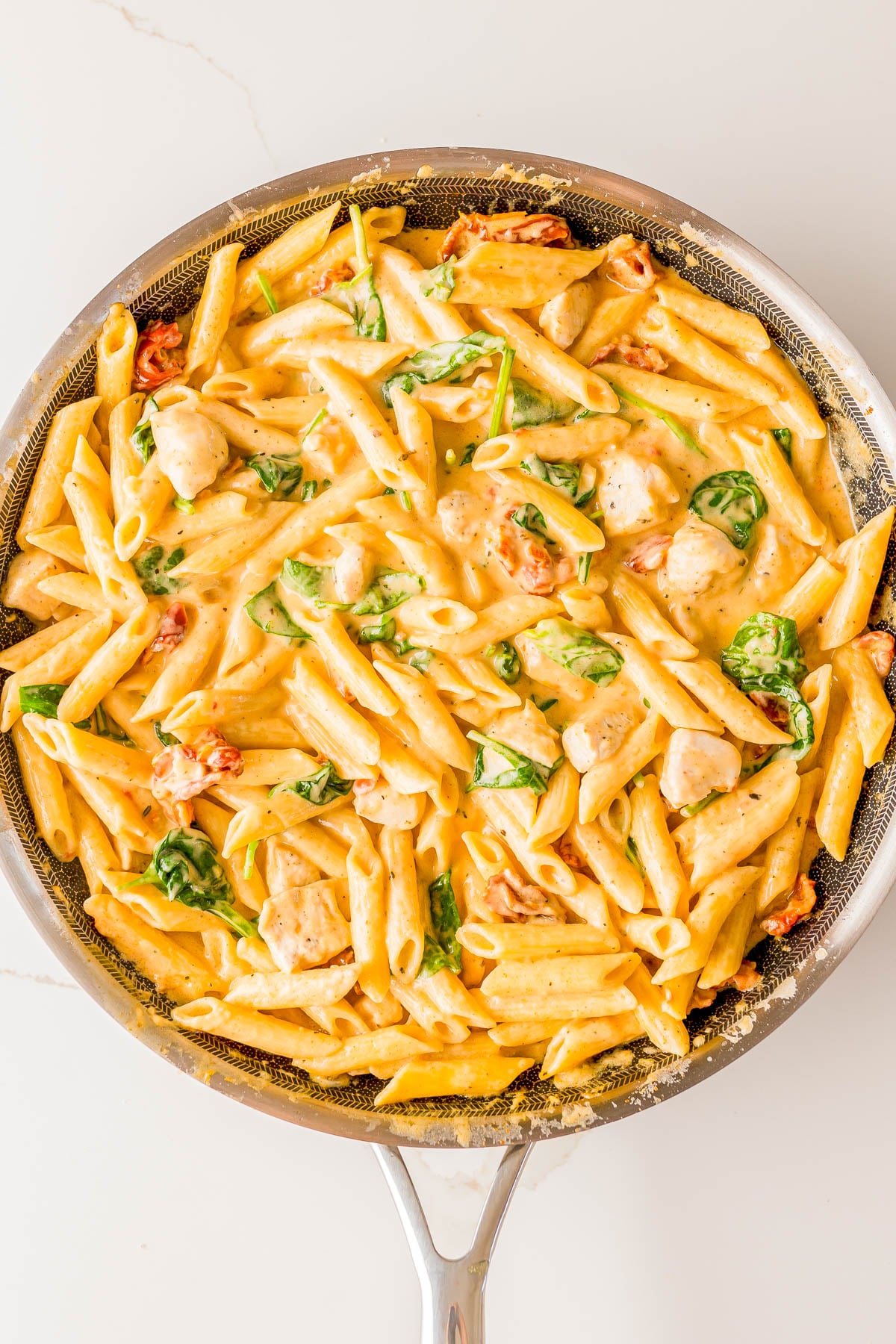 A skillet filled with creamy penne pasta, pieces of chicken, spinach, and sun-dried tomatoes.