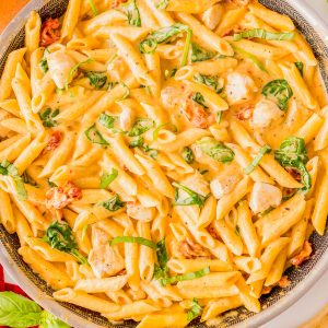 A skillet filled with creamy penne pasta, pieces of chicken, spinach, sun-dried tomatoes, and fresh basil, with a wooden spoon and basil leaves nearby.