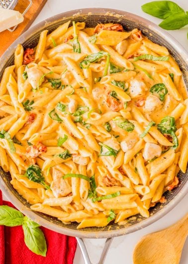 A skillet filled with creamy penne pasta, pieces of chicken, spinach, sun-dried tomatoes, and fresh basil, with a wooden spoon and basil leaves nearby.