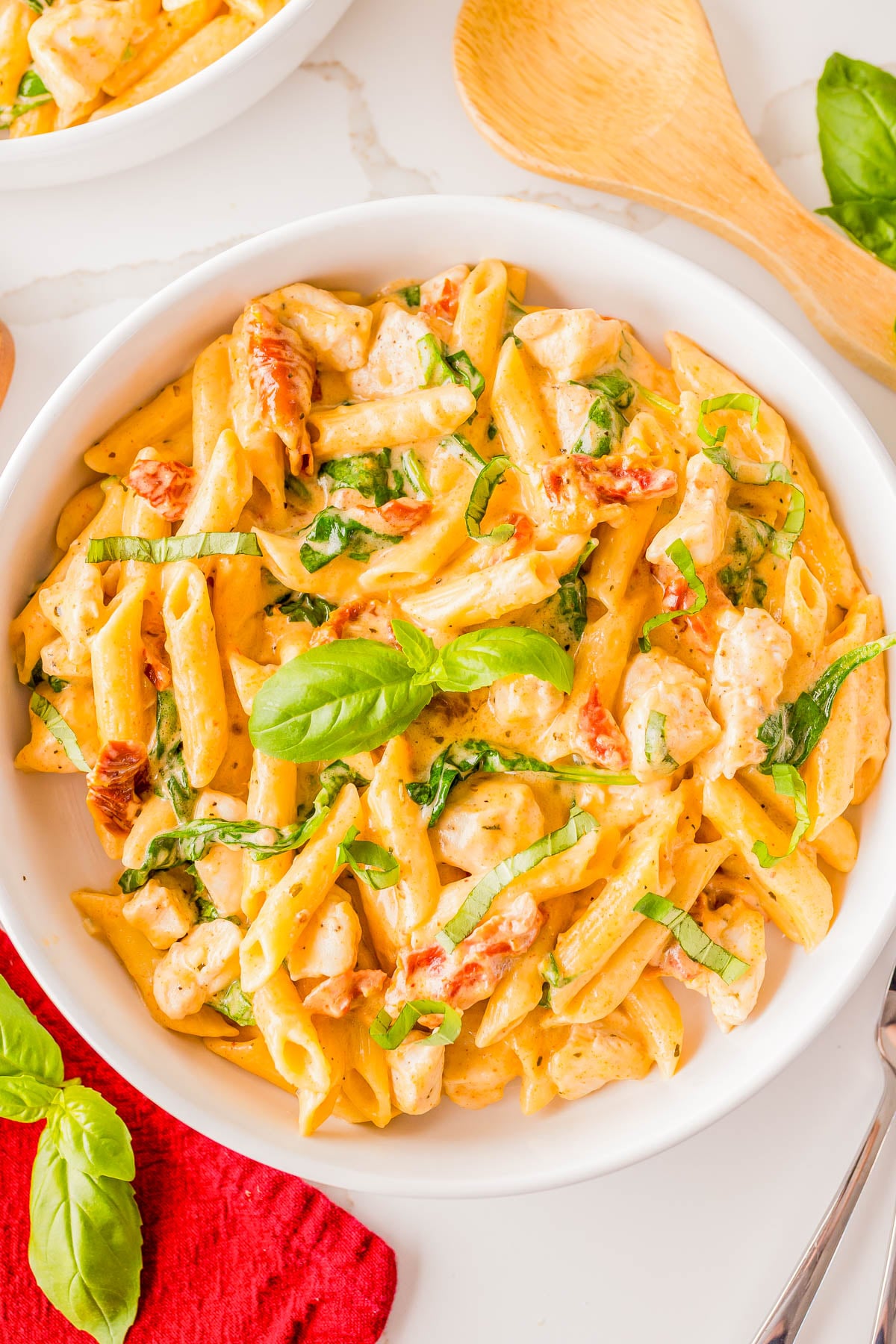 A bowl of creamy penne pasta with chicken, sun-dried tomatoes, spinach, and fresh basil, served with a wooden spoon and garnished with basil leaves.