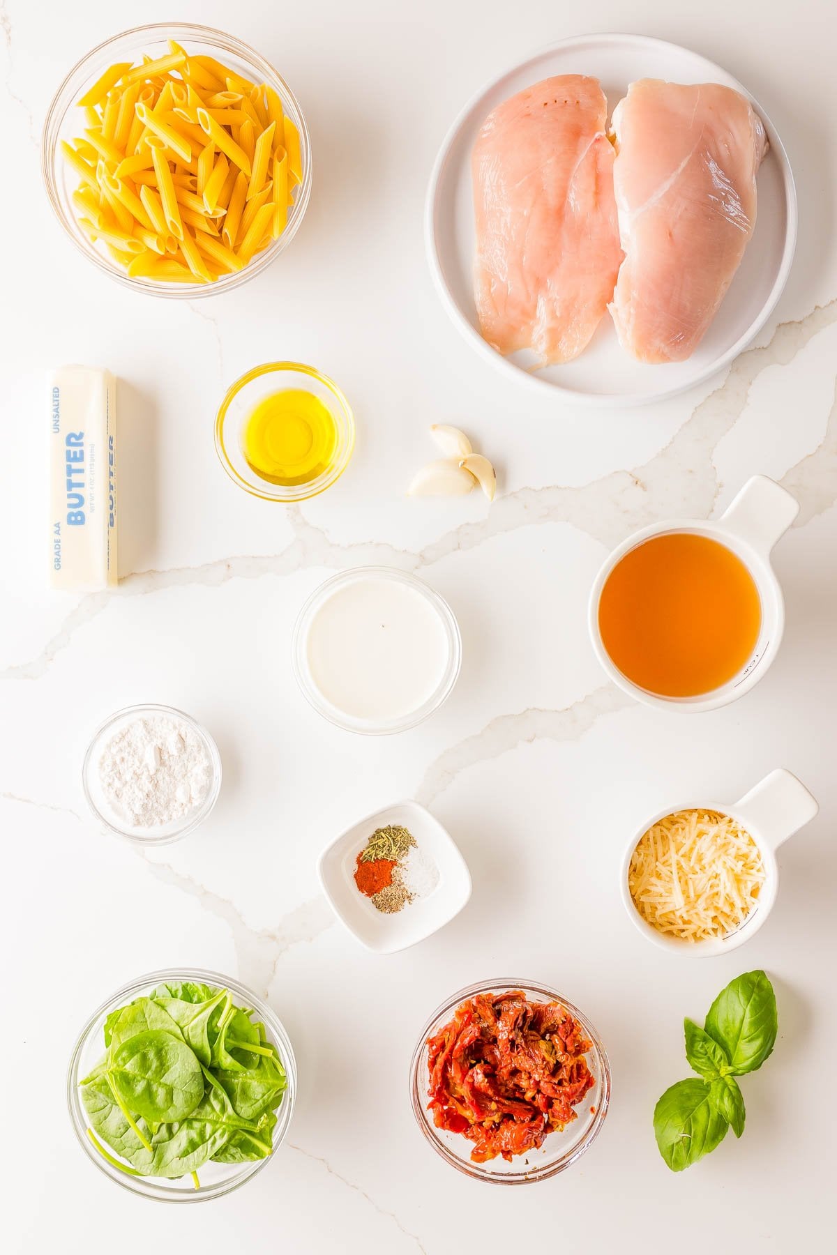 Ingredients for a pasta dish arranged on a white surface, including raw chicken breasts, pasta, butter, oil, garlic, cream, broth, flour, cheese, spinach, sun-dried tomatoes, and seasonings.