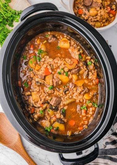 A slow cooker filled with beef and barley stew, containing chunks of beef, carrots, potatoes, and parsley, with a wooden spoon and a bowl of stew nearby.