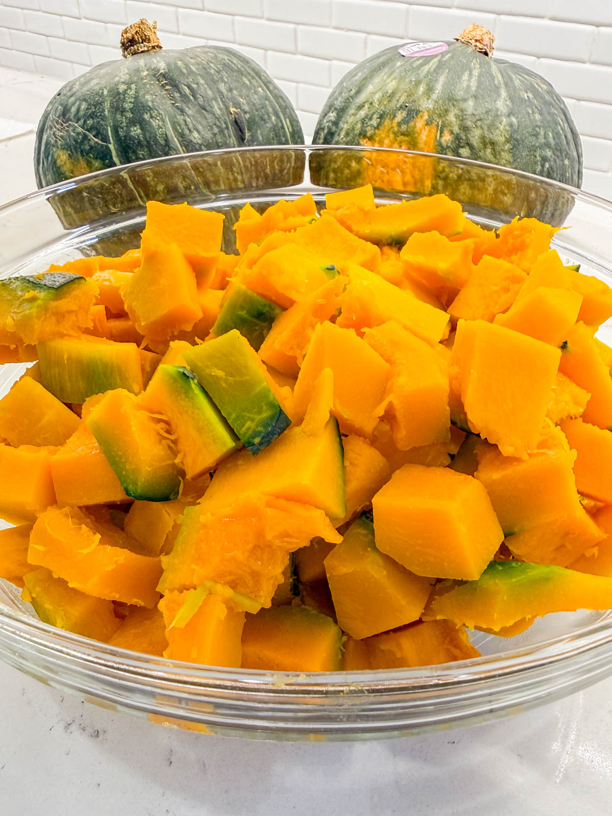 A glass bowl filled with cubed kabocha squash sits on a counter, with two whole kabocha squashes in the background.