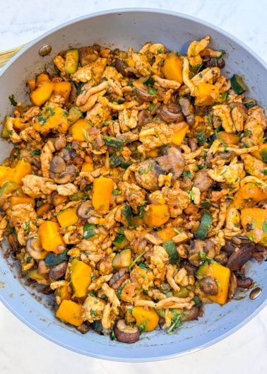A skillet filled with a cooked mixture of ground meat, diced butternut squash, zucchini, mushrooms, and herbs on a white surface.
