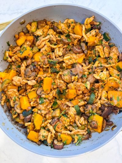 A skillet filled with a cooked mixture of ground meat, diced butternut squash, zucchini, mushrooms, and herbs on a white surface.
