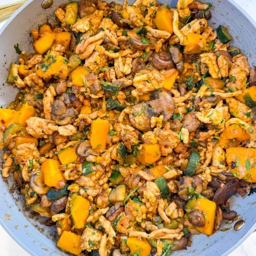 A skillet filled with a cooked mixture of ground meat, diced butternut squash, zucchini, mushrooms, and herbs on a white surface.