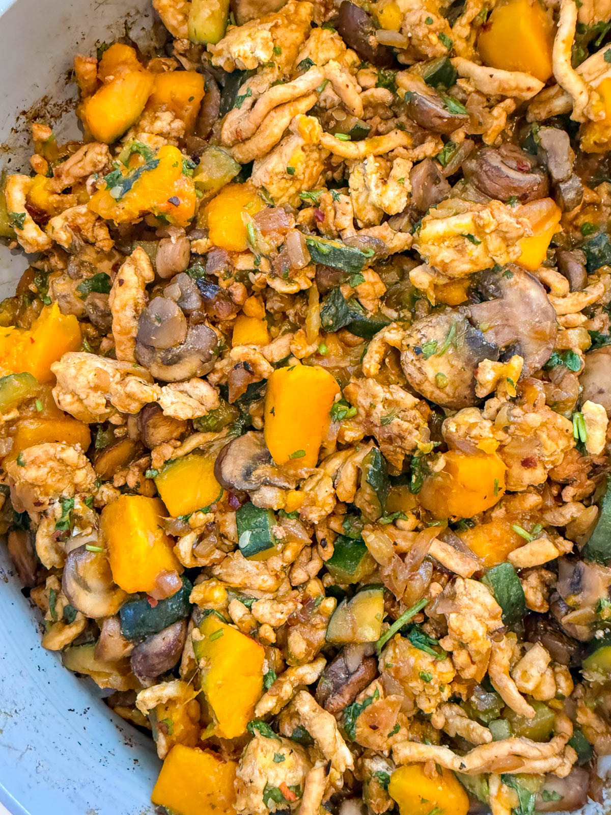 A close-up of a cooked dish with diced yellow squash, sliced mushrooms, ground meat, and green herbs mixed together.