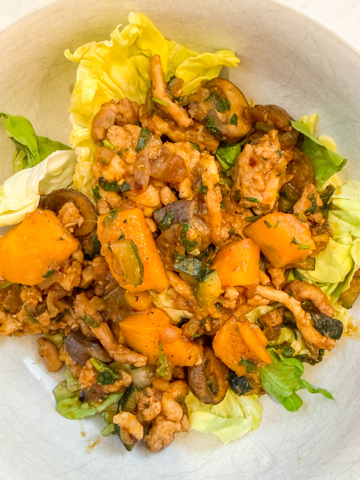 A bowl of cooked chicken pieces, yellow squash, zucchini, and eggplant served on a bed of lettuce leaves.