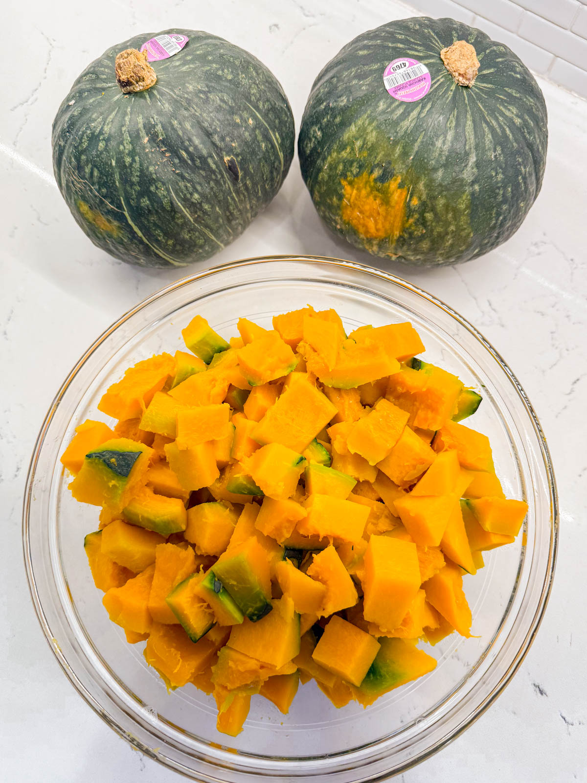 Two whole kabocha squash sit behind a glass bowl filled with peeled and cubed kabocha squash on a white countertop.
