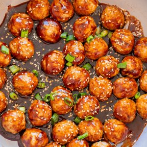 Meatballs coated in a dark glaze, garnished with sesame seeds and chopped green onions, arranged in a pan.