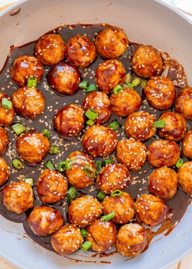 Meatballs coated in a dark glaze, garnished with sesame seeds and chopped green onions, arranged in a pan.