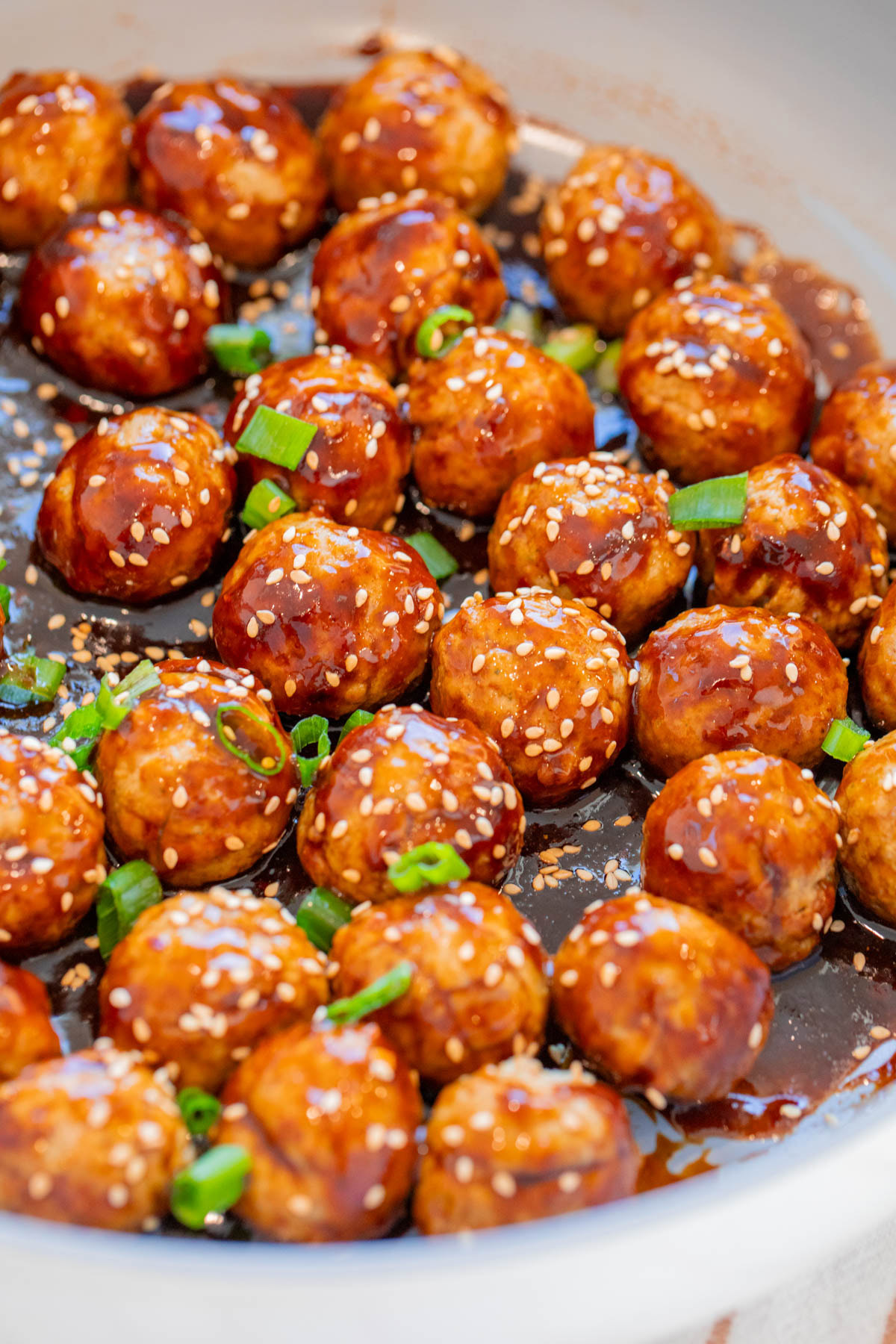 Meatballs coated in a glossy brown sauce, garnished with sesame seeds and chopped green onions, served in a shallow dish.