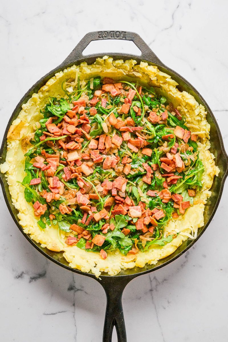 A cast iron skillet filled with mashed potatoes, chopped greens, and diced cooked bacon on a marble surface.