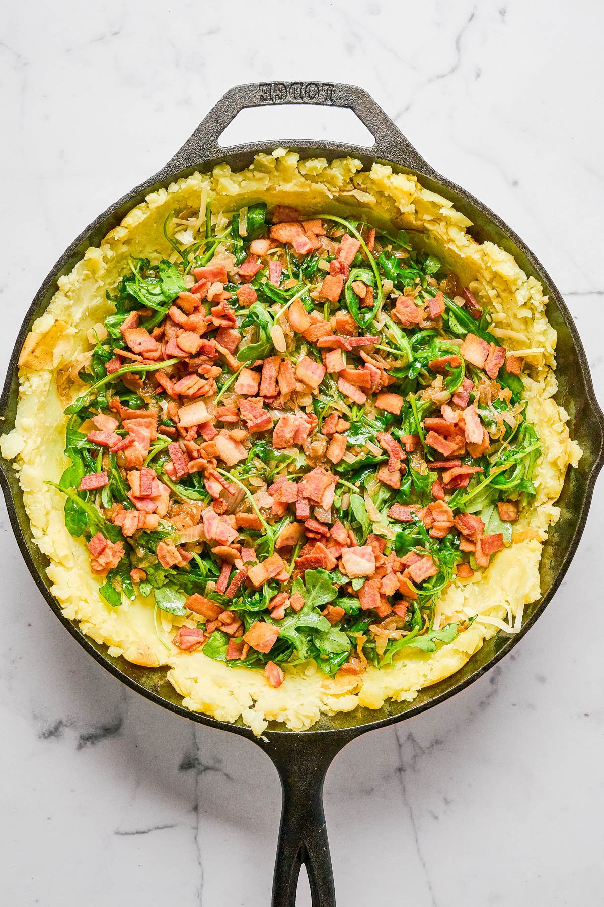A cast iron skillet filled with mashed potatoes, chopped greens, and diced cooked bacon on a marble surface.