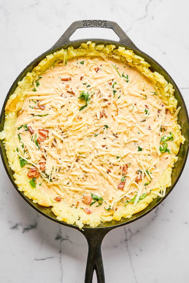 A cast iron skillet with a layer of mashed potatoes topped with a creamy mixture of cheese, herbs, and diced tomatoes on a marble surface.