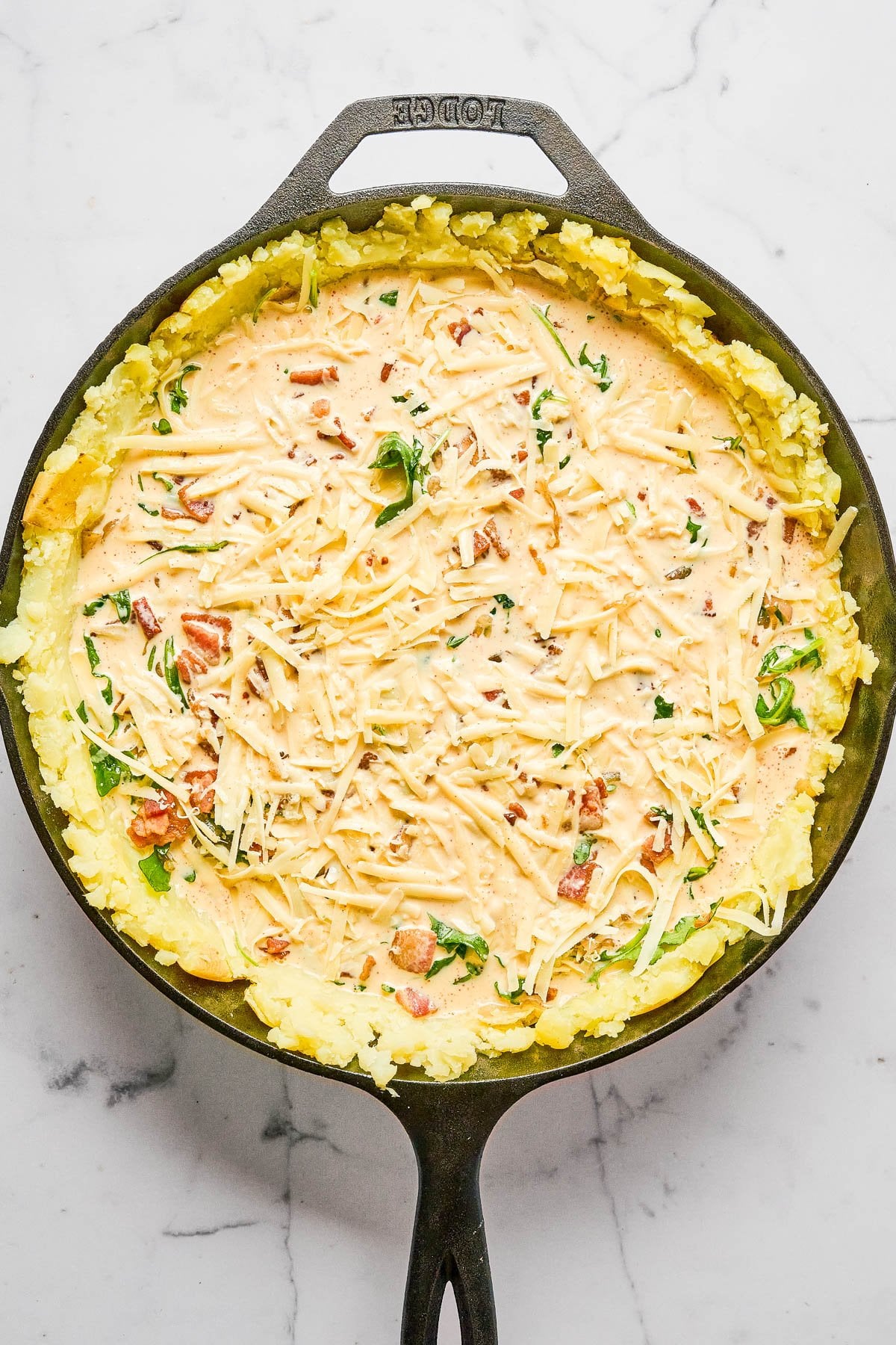 A cast iron skillet with a layer of mashed potatoes topped with a creamy mixture of cheese, herbs, and diced tomatoes on a marble surface.