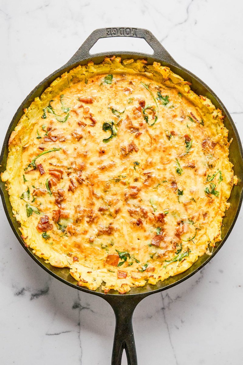 A baked frittata with visible spinach and pieces of ham in a cast iron skillet on a white marble surface.