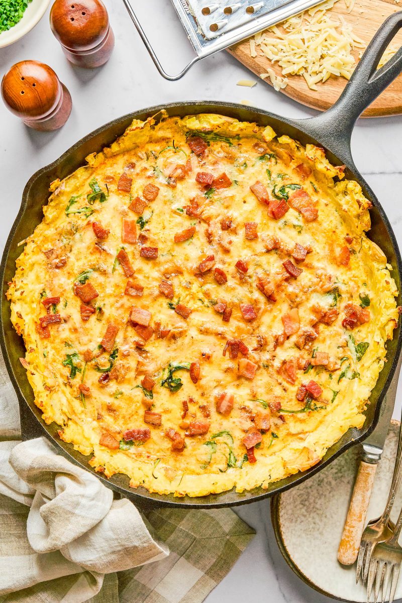 A cast iron skillet filled with a baked cheesy potato and bacon casserole sits on a table, surrounded by utensils, a napkin, and grated cheese.