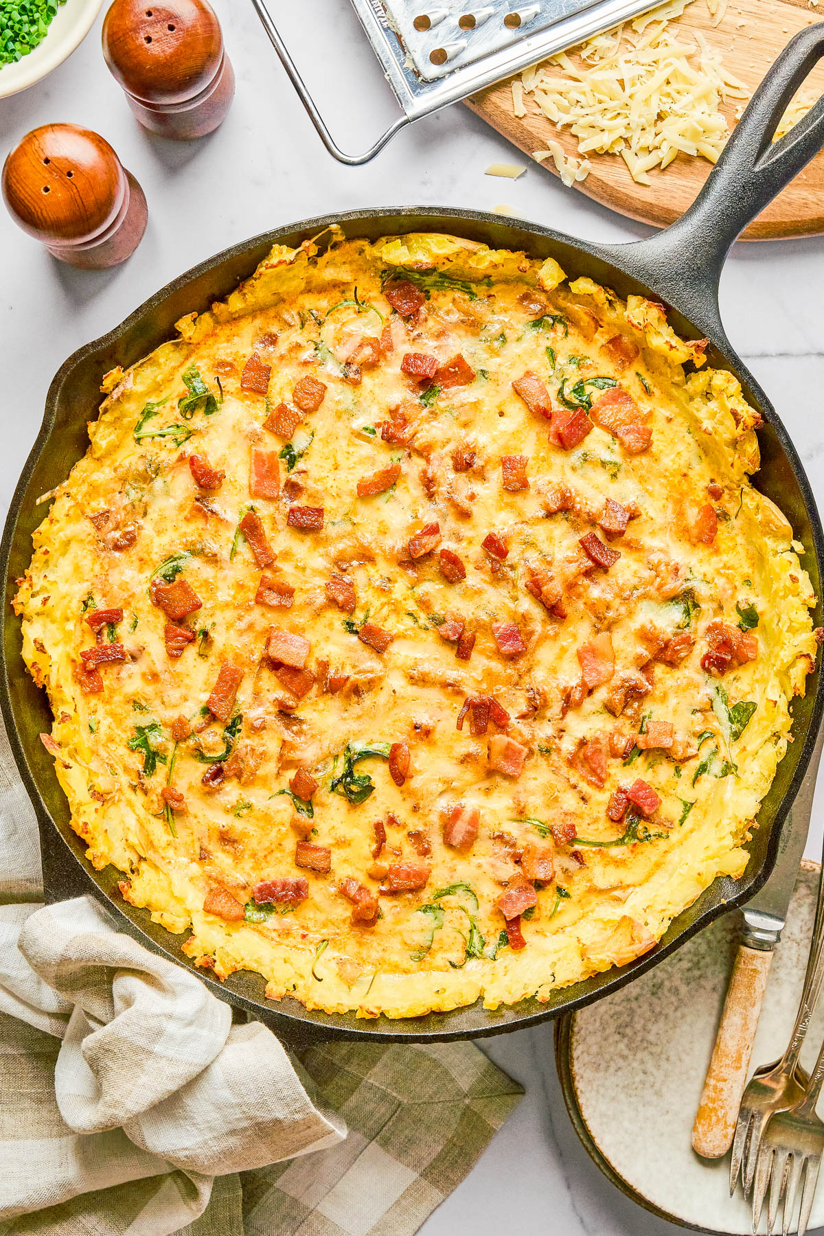 A cast iron skillet filled with a baked cheesy potato and bacon casserole sits on a table, surrounded by utensils, a napkin, and grated cheese.