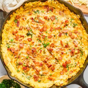 A skillet filled with a baked egg and potato dish topped with chopped bacon and herbs, with grated cheese, chives, and utensils nearby.