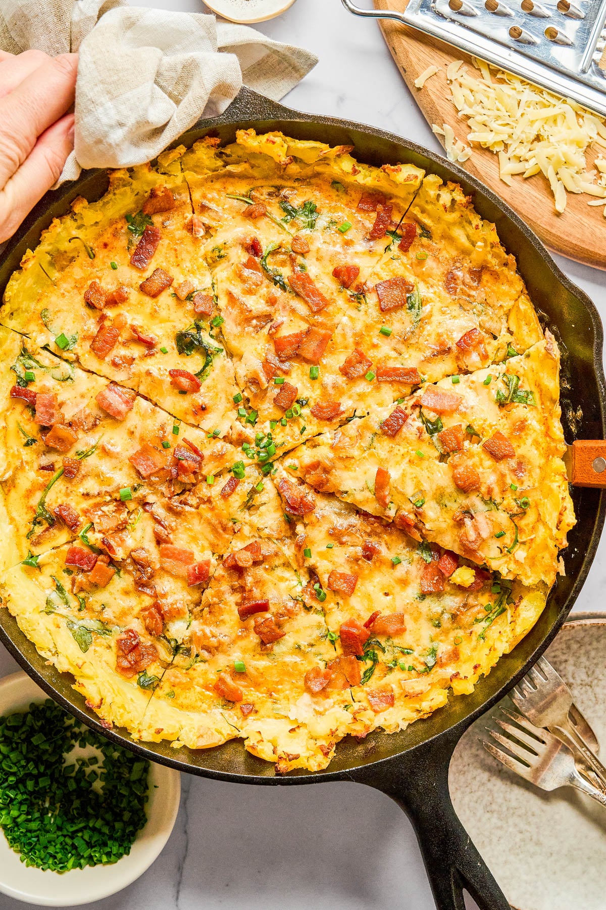 A sliced potato and bacon frittata in a cast iron skillet, topped with chopped herbs, with a grater, shredded cheese, and a bowl of chives nearby.