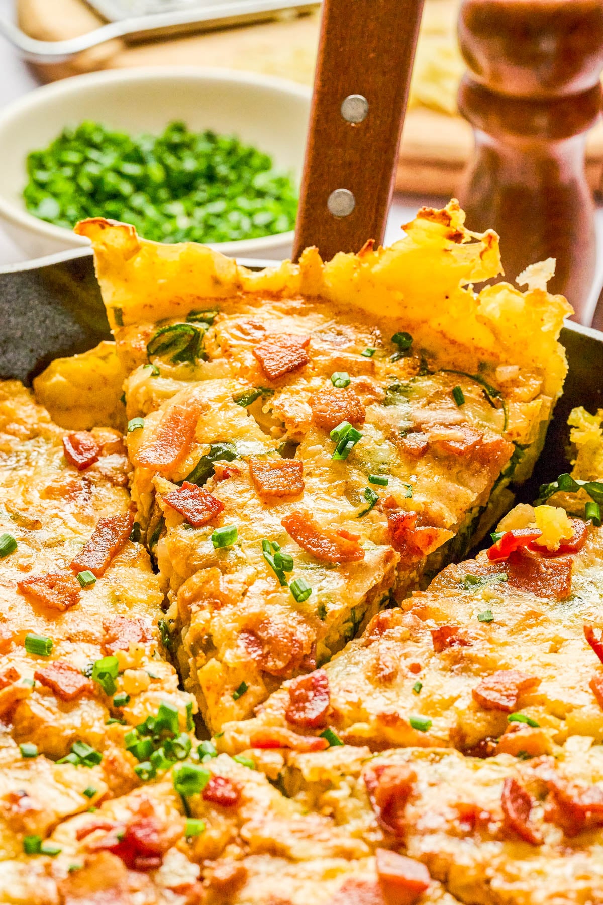 A close-up of a skillet quiche topped with chopped bacon and chives, with one slice being lifted out.