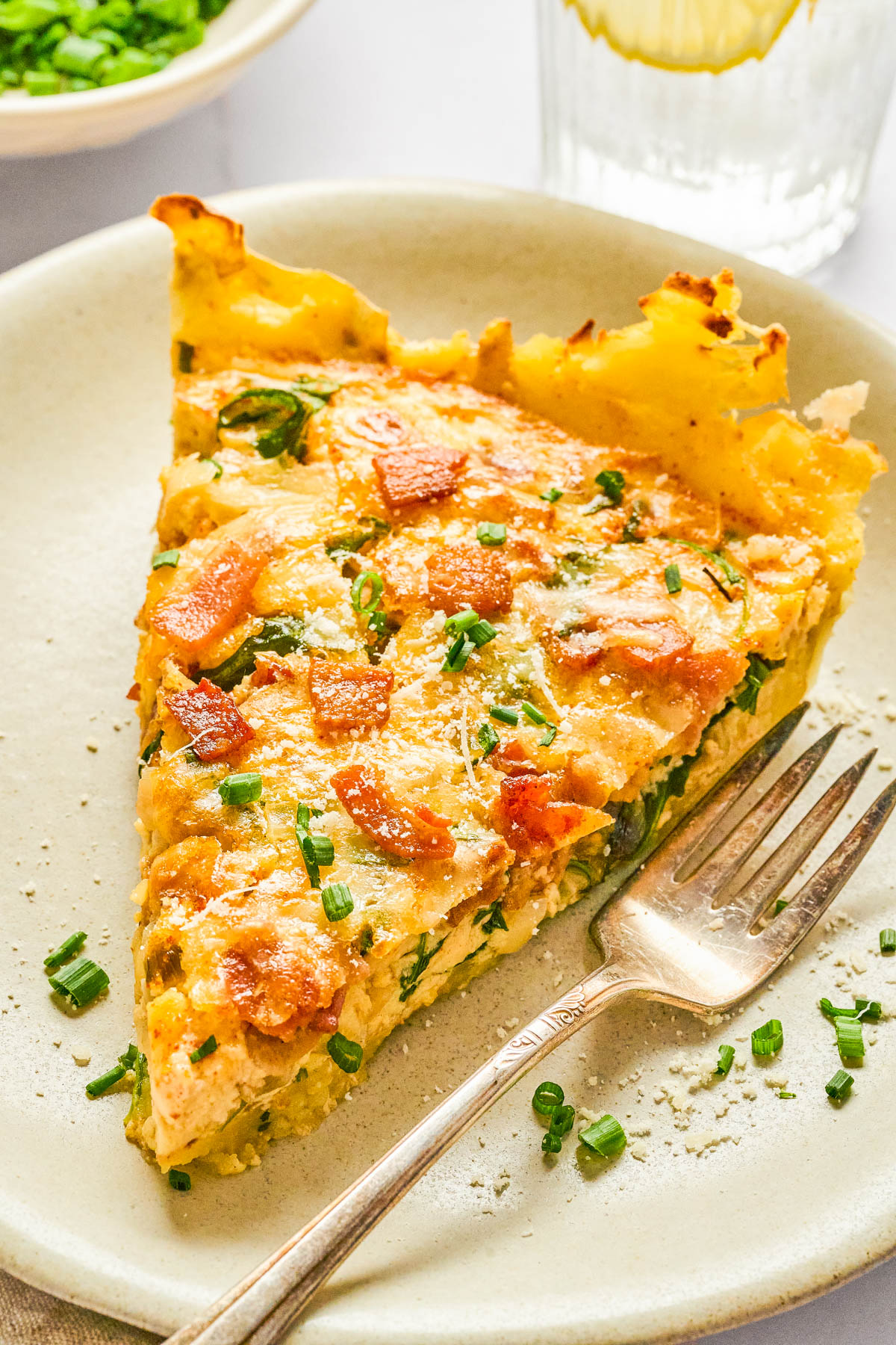 A slice of quiche topped with chopped chives and pieces of bacon, served on a beige plate with a fork.
