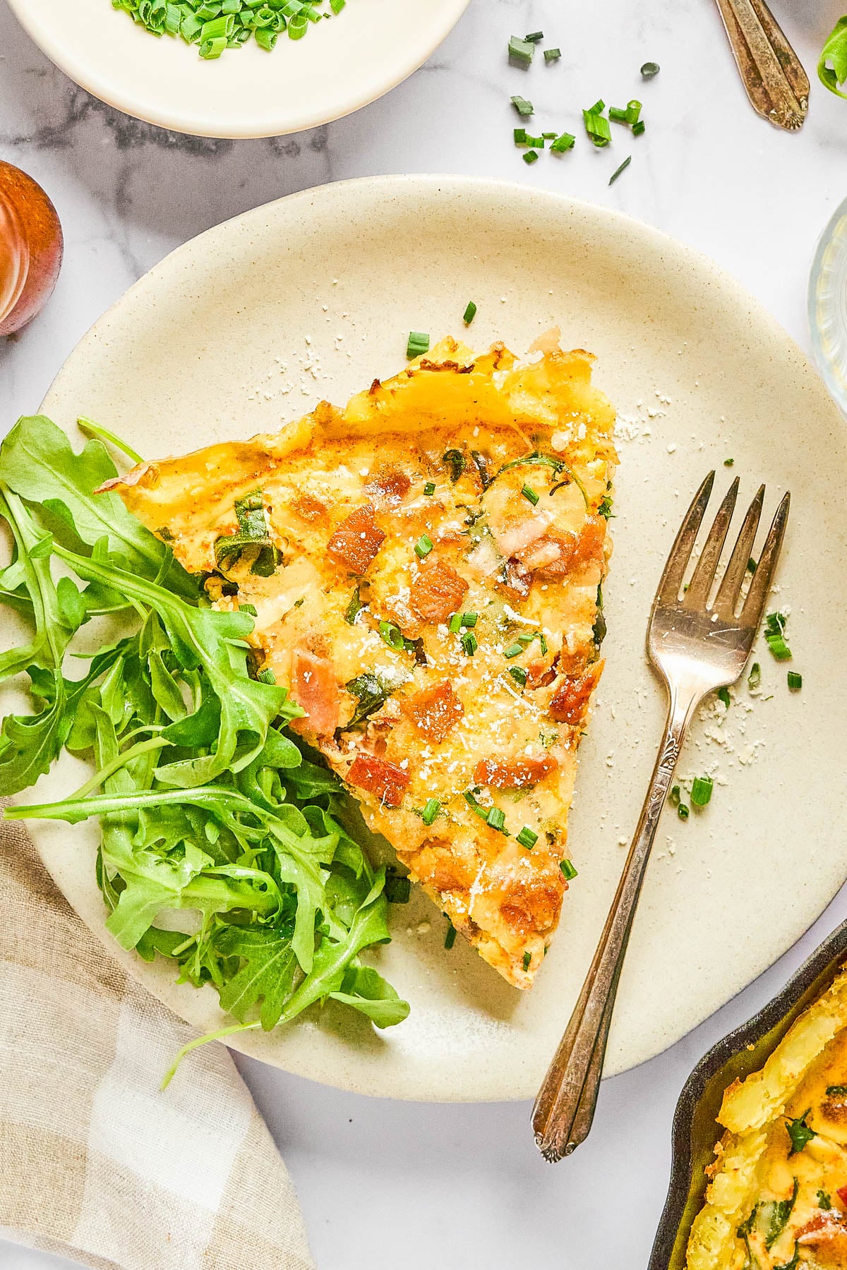 A slice of frittata with chopped chives and bacon is served on a plate with a side of arugula and a fork.