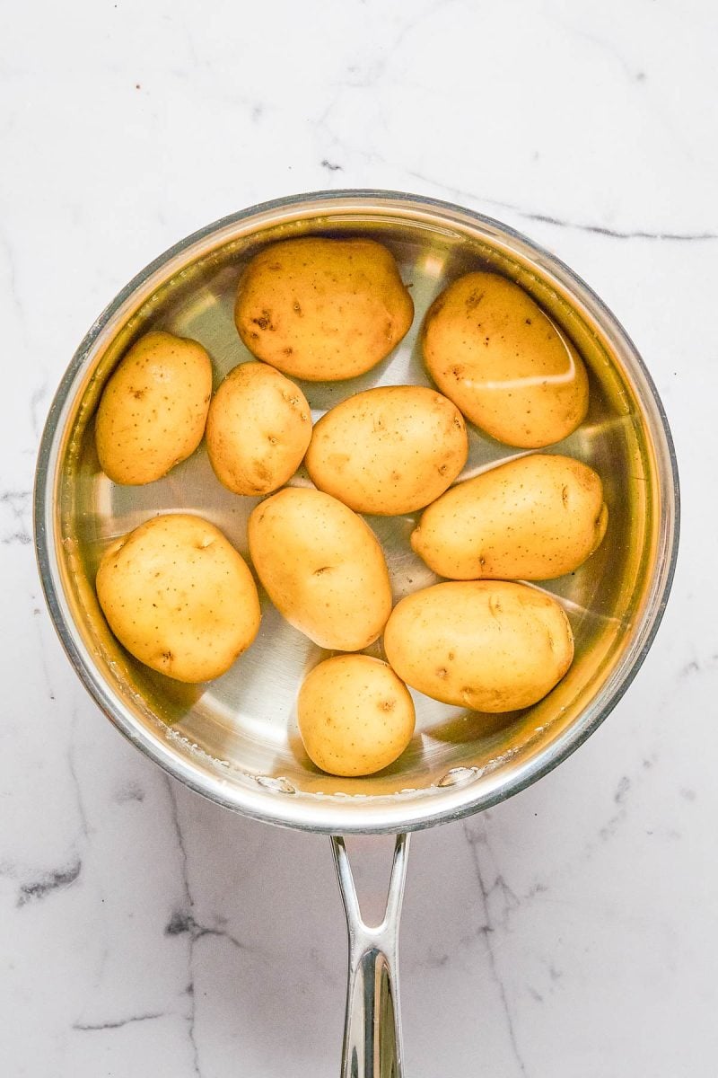 A stainless steel pot filled with whole yellow potatoes submerged in water, placed on a white marble surface.