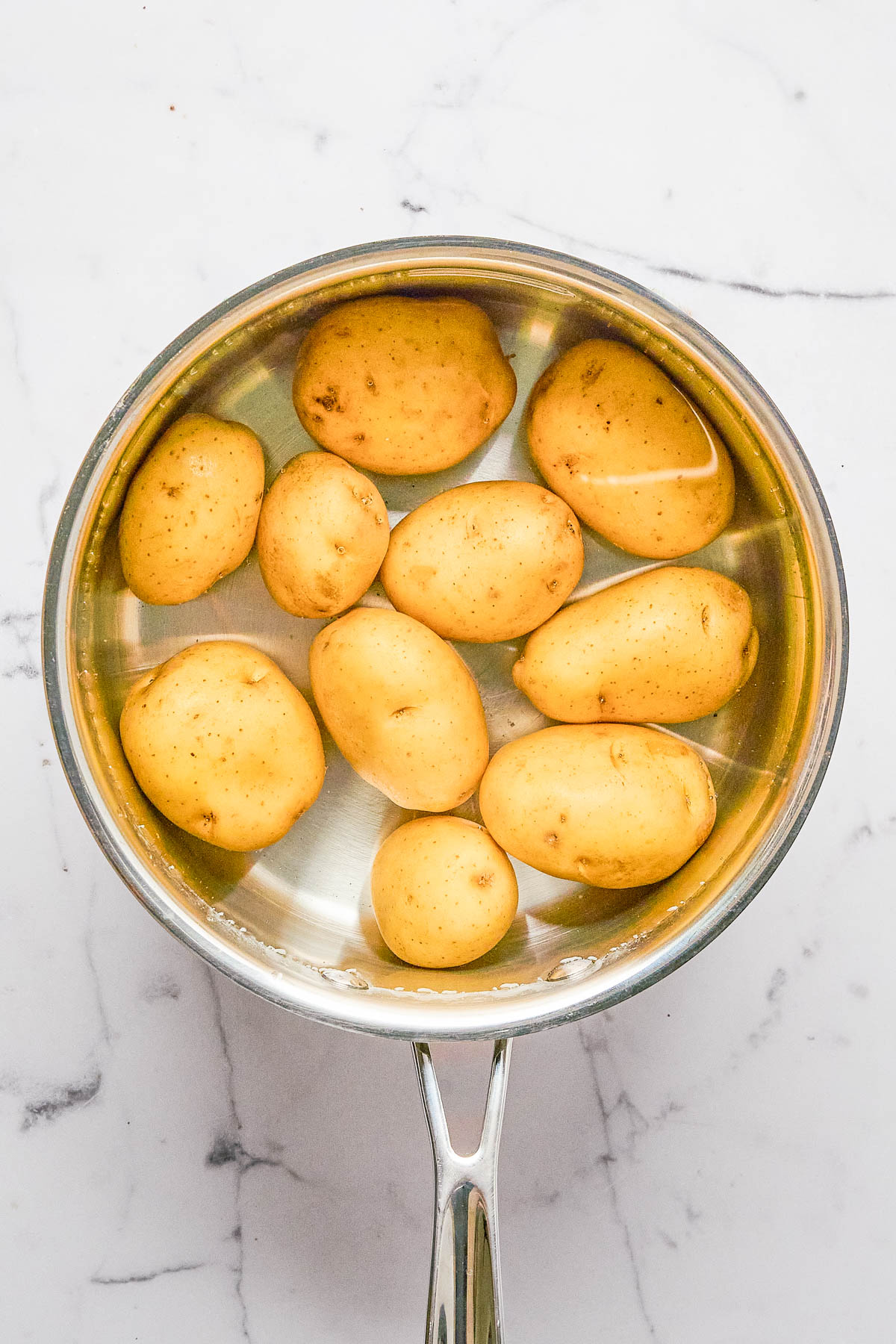 A stainless steel pot filled with whole yellow potatoes submerged in water, placed on a white marble surface.