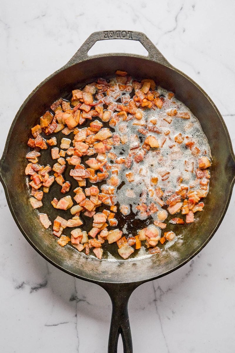 Cast iron skillet on a marble surface with diced bacon pieces partially cooked and sizzling in rendered fat.