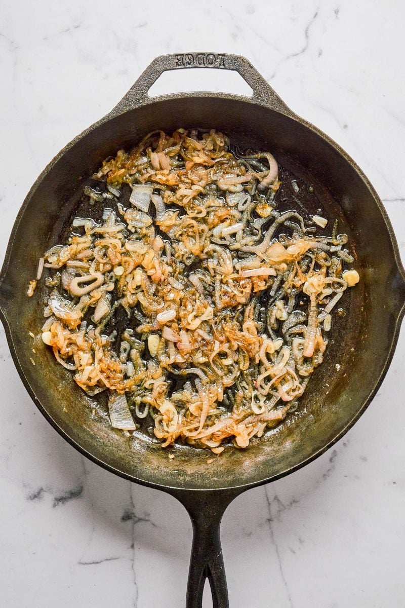 Sliced onions and shredded cheese are being sautéed in a black cast iron skillet on a white marble surface.