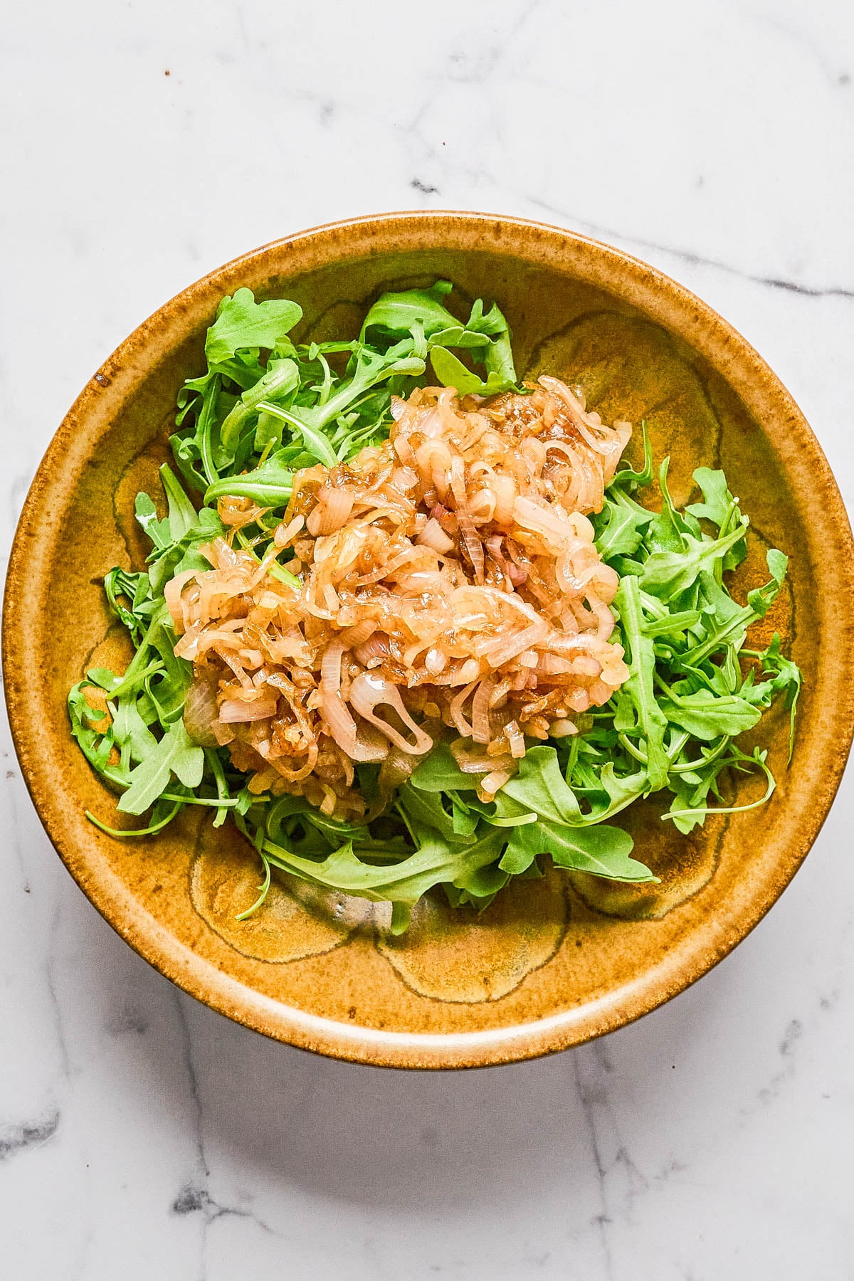 A brown bowl filled with fresh arugula topped with a portion of caramelized onions, set on a white marble surface.