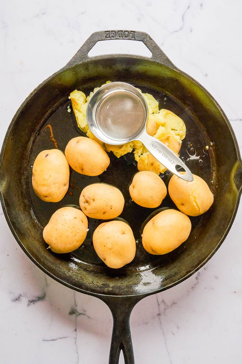 A cast iron skillet with whole yellow potatoes, some partially smashed with a metal measuring cup, on a white marble surface.