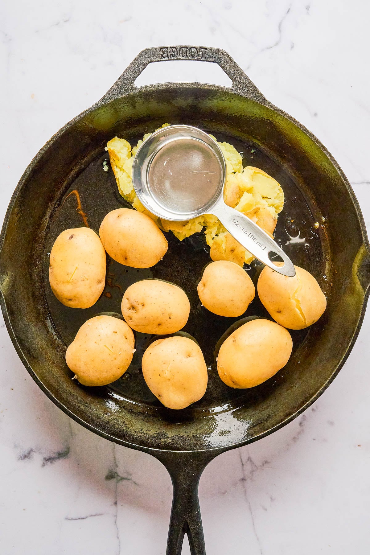 A cast iron skillet with whole yellow potatoes, some partially smashed with a metal measuring cup, on a white marble surface.