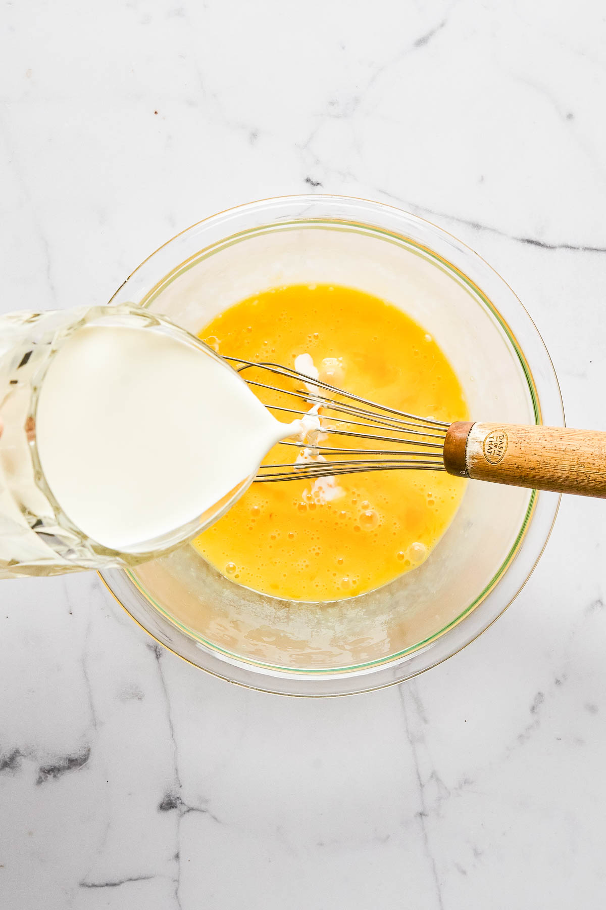 A glass bowl with beaten eggs is being whisked while milk is poured in from a glass, on a white marble countertop.