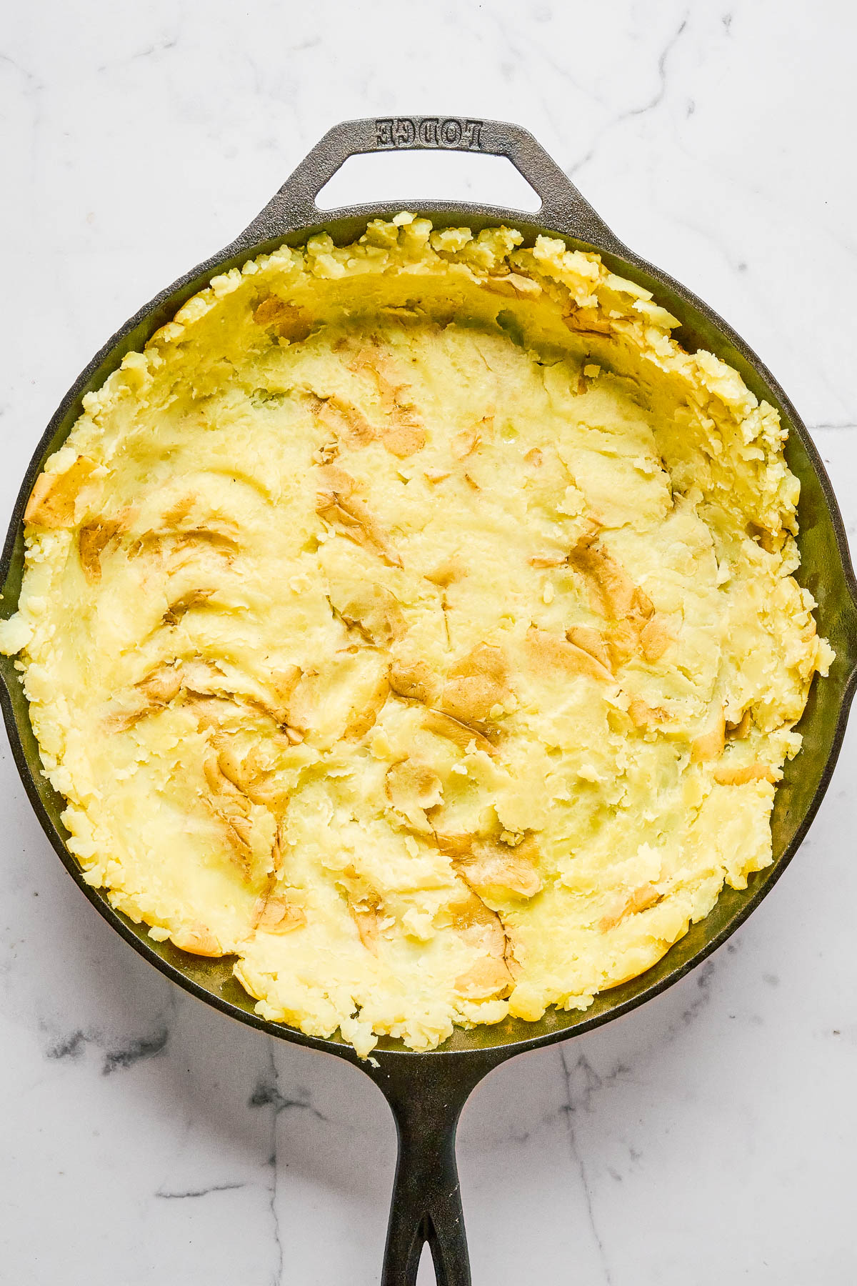 A cast iron skillet filled with mashed potatoes, spread evenly and lightly browned on top, sits on a white marble surface.